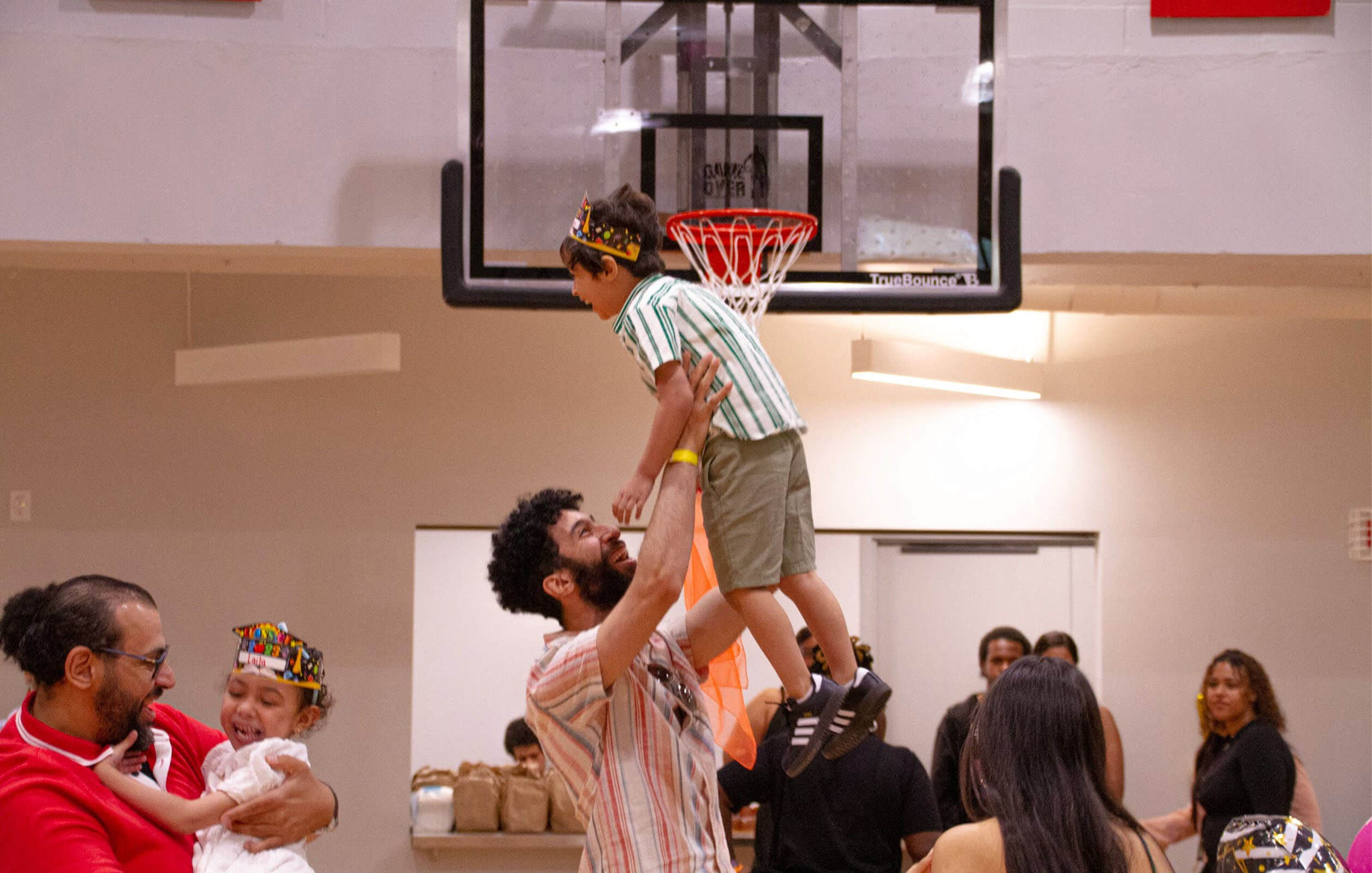 A man holding up a child and smiling on a basketball court inside a Grand Street Settlement center
